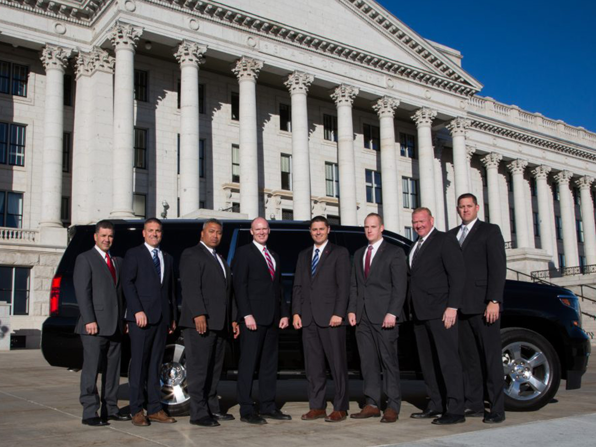 Eight members of the executive protection squad are wearing suits and standing in front of a a black SUV parked in front of the capitol.